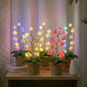 Op de afbeelding: Een verzameling van vijf gehaakte bloemenlampen in kleine potten. De bloemen zijn in tinten geel, roze en wit, met groene stelen en bladeren. Elke bloem heeft een klein lichtje binnenin, wat een zachte gloed creëert.