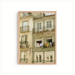 Może przedstawiać: Oprawiony wydruk fasady budynku z wieloma oknami i balkonami. Budynek ma jasny zielony i beżowy schemat kolorów. Pranie wisi z okna. Obraz jest oprawiony w jasnobrązową drewnianą ramę.