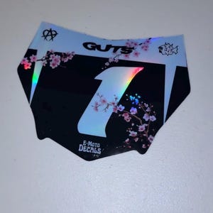 May include: Black and white motorcycle decal with a floral design and the word "GUTS". The number "1" is in the center with a rainbow holographic effect. The decal also has the text "E-MOTO DECALS".