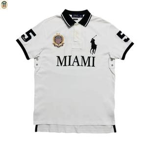 Puede incluir: Polo blanco con detalles negros, con la palabra "MIAMI" en negro. La camisa tiene un cuello blanco y negro, el número "5" en las mangas y un logotipo de jugador de polo.