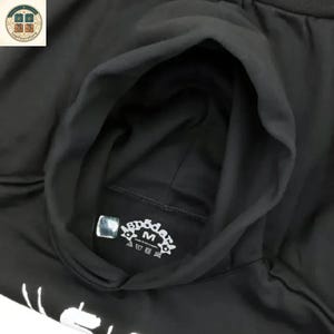 Puede incluir: Sudadera negra con capucha y una etiqueta con el texto "sp5der" y la letra "M". La sudadera con capucha est&aacute; hecha de un material suave.