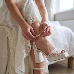 May include: White platform sandals with a square toe and block heel. The sandals feature a delicate beaded design on the straps and a buckle closure. The person is wearing a white lace dress. The sandals are a bridal or special occasion shoe.