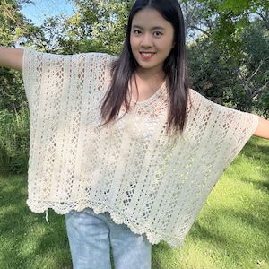 Pu&ograve; includere: Un poncho all'uncinetto color bianco sporco dal taglio ampio, caratterizzato da uno scollo a V e da una combinazione di motivi a righe verticali e dettagli in pizzo traforato. Il capo presenta un orlo smerlato ed &egrave; indossato sopra un top bianco e jeans azzurri.