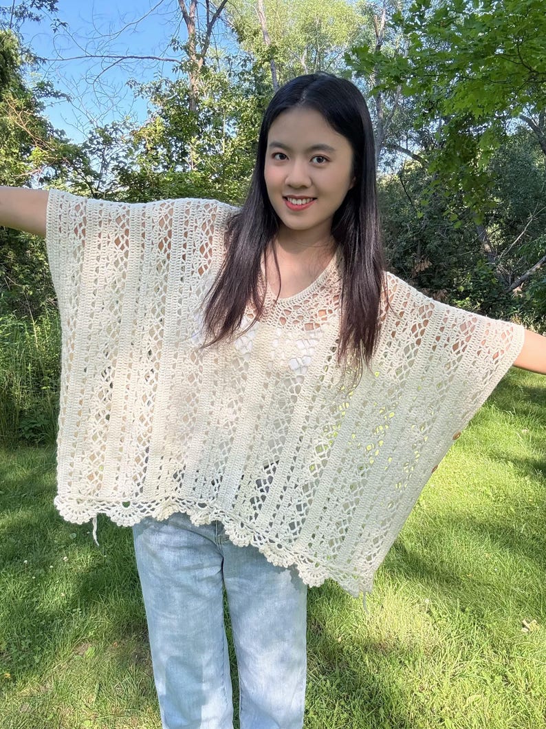 Pu&ograve; includere: Un poncho all'uncinetto color bianco sporco dal taglio ampio, caratterizzato da uno scollo a V e da una combinazione di motivi a righe verticali e dettagli in pizzo traforato. Il capo presenta un orlo smerlato ed &egrave; indossato sopra un top bianco e jeans azzurri.