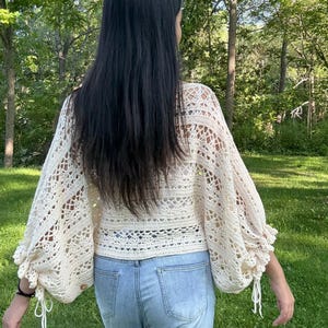 Pu&ograve; includere: Un top all'uncinetto color crema caratterizzato da lunghe e voluminose maniche a vescovo con lacci ai polsini e un complesso motivo traforato su tutto il corpetto e le maniche, abbinato a jeans in denim azzurro.