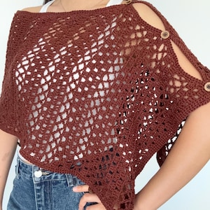 May include: A dark brown crochet poncho featuring an open diamond-shaped stitch pattern. The garment has a wide neckline and a series of small, round, light brown buttons along the shoulder seam, creating a cutout effect on the upper arm area.