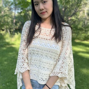 Pu&ograve; includere: Un poncho all'uncinetto color crema caratterizzato da un intricato motivo traforato con varie fasce orizzontali di punti simili al pizzo, un ampio scollo a barchetta e bordi smerlati che ricadono morbidamente sulle spalle e sul busto.