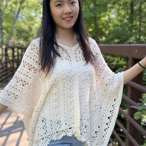 Pu&ograve; includere: Un poncho all'uncinetto color crema caratterizzato da un intricato motivo in pizzo traforato con scollo a V e orlo smerlato. Il capo &egrave; indossato sopra un top bianco e jeans azzurri, mostrando una silhouette ampia e fluida con maniche larghe.