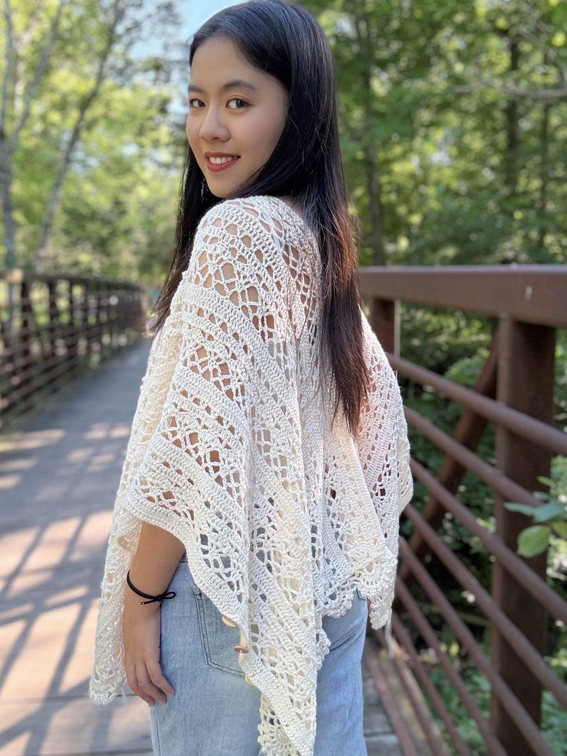Pu&ograve; includere: Un poncho all'uncinetto color bianco sporco caratterizzato da un intricato motivo traforato con disegni geometrici e floreali. Il capo presenta una silhouette ampia e drappeggiata con orli a punta e piccoli bottoni in legno sui lati.