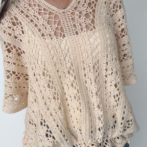 Puede incluir: Top de crochet color crema con cuello en V y mangas tres cuartos. El diseño calado presenta un delicado patrón en toda la prenda. El dobladillo inferior tiene un borde festoneado. La parte superior se usa sobre una prenda interior de color más claro.