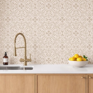 May include: A kitchen scene featuring a wall with a beige and white damask pattern, a brass gooseneck faucet, a dark brown soap dispenser, and a white bowl filled with yellow lemons on a white countertop above light wood cabinets.