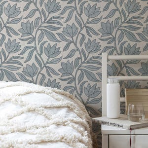 May include: A bedroom scene featuring a white bed with a textured, fringed throw. The wall is adorned with a floral wallpaper in shades of blue and white. A white water bottle and a glass of water sit on a nightstand.