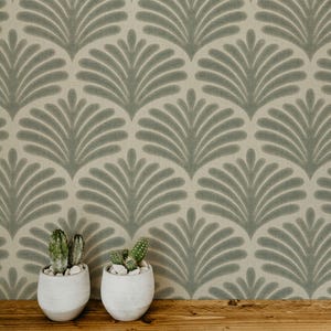 May include: Two small green cacti in white ceramic pots sit in front of a wallpaper featuring a repeating sage green fan-shaped leaf pattern on a light beige background. The plants have visible spines and are potted in simple round containers.