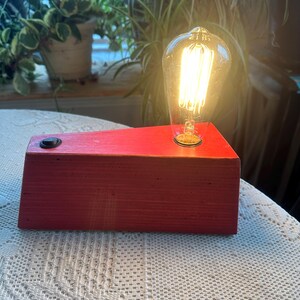 May include: A red wooden desk lamp with a vintage-style Edison bulb. The lamp has a rectangular base with a black on/off switch. The bulb is clear with visible filaments, emitting a warm glow. The lamp is on a white lace tablecloth.
