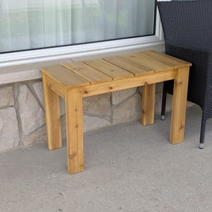 May include: A small, rectangular wooden bench with a natural finish. The bench has a slatted top and four sturdy legs. It is approximately 30 inches long, 12 inches wide, and 18 inches high. The bench is made of light-colored wood.