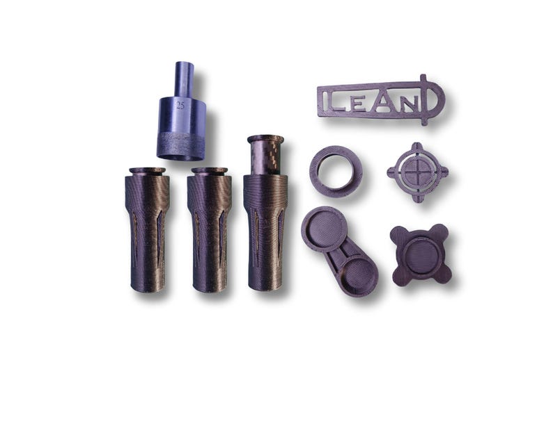 May include: An assortment of dark gray metal machine parts, including cylindrical pieces, a ring, and a component with the word "LEAN" cut out. One part has the number "25" etched on it. The parts are arranged on a white background.
