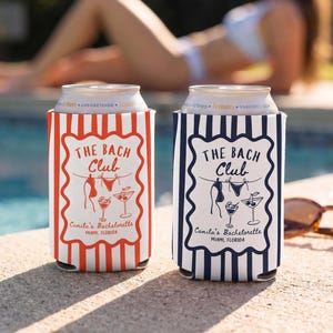 May include: Two cylindrical beverage can coolers featuring vertical stripes and a central graphic with the text THE BACH Club, Camila's Bachelorette, MIAMI, FLORIDA, and illustrations of swimwear and cocktails. One cooler is orange, the other is navy blue.