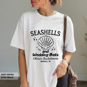 以下が含まれることがあります： 白地のTシャツ。黒いグラフィックで「SEASHELLS and Wedding Bells Olivia's Bachelorette NASHVILLE, TN」と書かれています。貝殻のイラストと「est. 2026」の文字も含まれています。