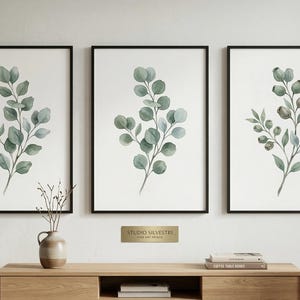 Könnte beinhalten: Drei gerahmte botanische Drucke mit Aquarellillustrationen von Eukalyptuszweigen in Grün- und Blau-Grau-Tönen. Die Drucke werden über einem Holzschrank ausgestellt. Die Rahmen sind schwarz.