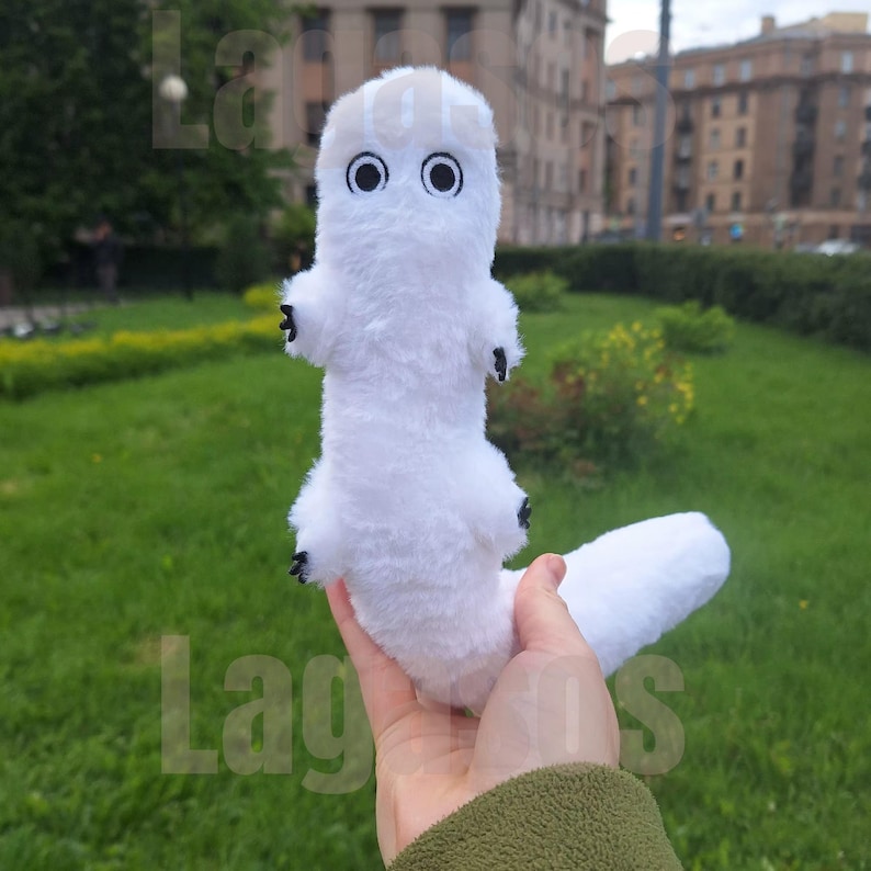 May include: A white plush toy with a long, cylindrical body, a rounded head, and a long tail. It features two large, circular black eyes and small black claws on its four limbs. The texture of the plush is soft and fuzzy, with a light beige patch on top.