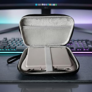 May include: Open black zippered case with a light gray interior, holding a light gray handheld gaming device. The case has a mesh pocket and a strap to secure the device. The case is on a dark surface.