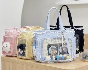 May include: A display of pastel-colored tote bags with clear front pockets. The bags are in pink, yellow, light blue, and black, each with lace trim and a top handle. The light blue bag has a pearl and charm embellishment. The bags contain plush toys and badges.