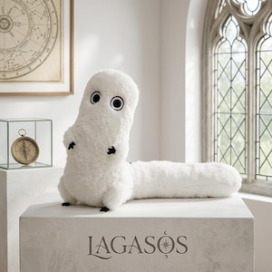 May include: A white plush toy with a long, cylindrical body and a raised head. It features two large, circular black and white eyes and small black appendages. The base of the display features the text LAGASOS with a small star symbol above the O.