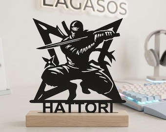 May include: A black metal silhouette of a crouching ninja holding a sword, set against a star-shaped background. The base is a rectangular piece of light-colored wood. The word HATTORI is cut out in capital letters at the bottom of the design.