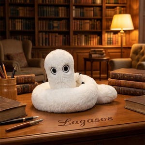 May include: A white plush toy with a coiled body and two large black-rimmed eyes sits on a wooden desk. The desk surface features the engraved text Lagasos, surrounded by stacks of antique books, a wooden pen holder, and two fountain pens.