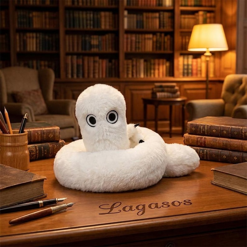 May include: A white plush toy with a coiled body and two large black-rimmed eyes sits on a wooden desk. The desk surface features the engraved text Lagasos, surrounded by stacks of antique books, a wooden pen holder, and two fountain pens.