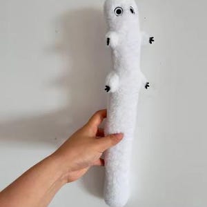 May include: A long, white, plush stuffed animal with a soft, fuzzy texture. It features a cylindrical body, two small arms with black-tipped fingers, and two round, black-and-white eyes positioned near the top of its head.