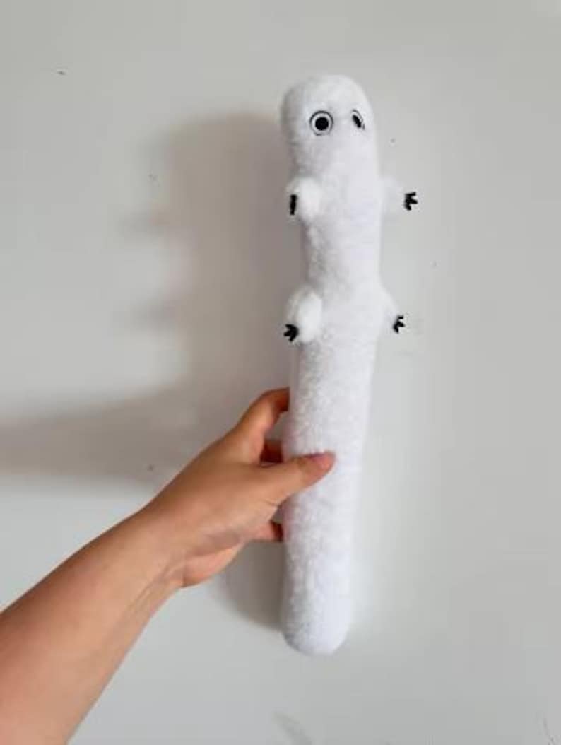 May include: A long, white, plush stuffed animal with a soft, fuzzy texture. It features a cylindrical body, two small arms with black-tipped fingers, and two round, black-and-white eyes positioned near the top of its head.
