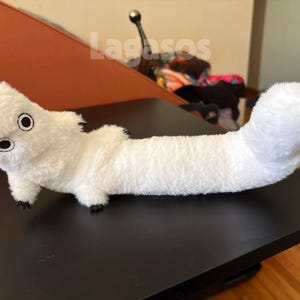 May include: A long, white plush toy with a soft, fuzzy texture, featuring a rounded head with two large, circular black and white eyes, small limbs, and a thick, curved tail. The word Lagasos is printed in white letters above the plush toy.