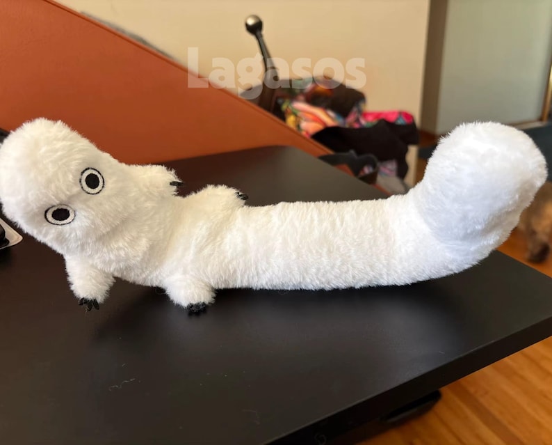 May include: A long, white plush toy with a soft, fuzzy texture, featuring a rounded head with two large, circular black and white eyes, small limbs, and a thick, curved tail. The word Lagasos is printed in white letters above the plush toy.