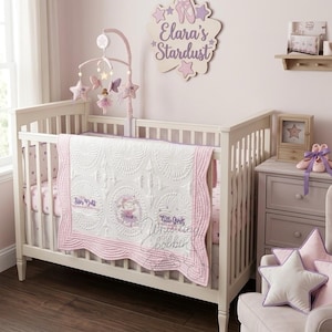 May include: A white crib with a pink and white quilt and bedding set. The quilt features a ballerina design and the words "Star Doll" and "Little Girls Whistling bobbin". A mobile with stars and fairies hangs above the crib. A wooden sign says "Elara's Stardust".