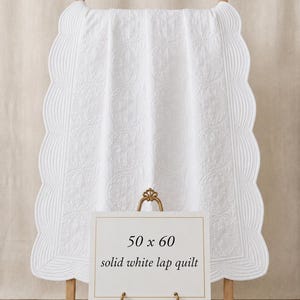 May include: A solid white lap quilt featuring a scalloped edge and intricate circular quilted patterns. A sign in front displays the text 50 x 60 solid white lap quilt. The quilt is draped over a wooden stand, showcasing its texture and design.