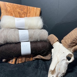May include: A stack of three yarn skeins in cream, gray, and brown, labeled "RAM". The yarn is displayed on a wooden base, with a ram skull and horns in the background. The yarn is fluffy and appears to be a natural fiber.