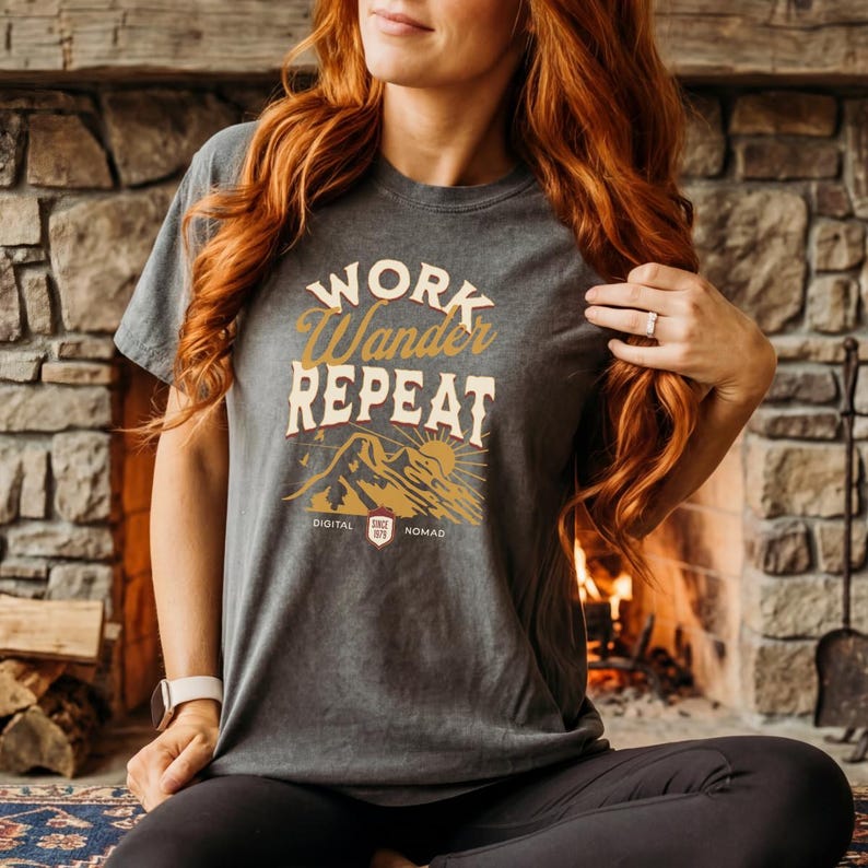 May include: Dark gray t-shirt with the words "WORK Wander REPEAT" in a gold and white design. The graphic includes a mountain scene with a sun and the text "DIGITAL NOMAD" below. The shirt is a casual style.