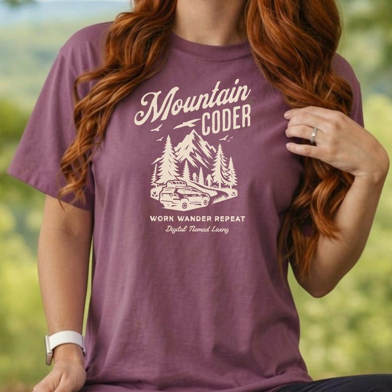 Może przedstawiać: Fioletowa koszulka z kr&oacute;tkim rękawem z napisem "Mountain Coder" w stylizowanej czcionce. Poniżej tekstu znajduje się grafika samochodu w g&oacute;rskim krajobrazie. Poniżej grafiki znajduje się tekst "Work Wander Repeat Digital Nomad Living".