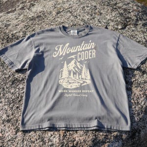 Może przedstawiać: Szara koszulka z nadrukiem graficznym. Projekt zawiera słowa "Mountain Coder" nad sceną g&oacute;rską i leśną. Poniżej widnieją słowa "Work Wander Repeat" i "Digital Nomad Living".