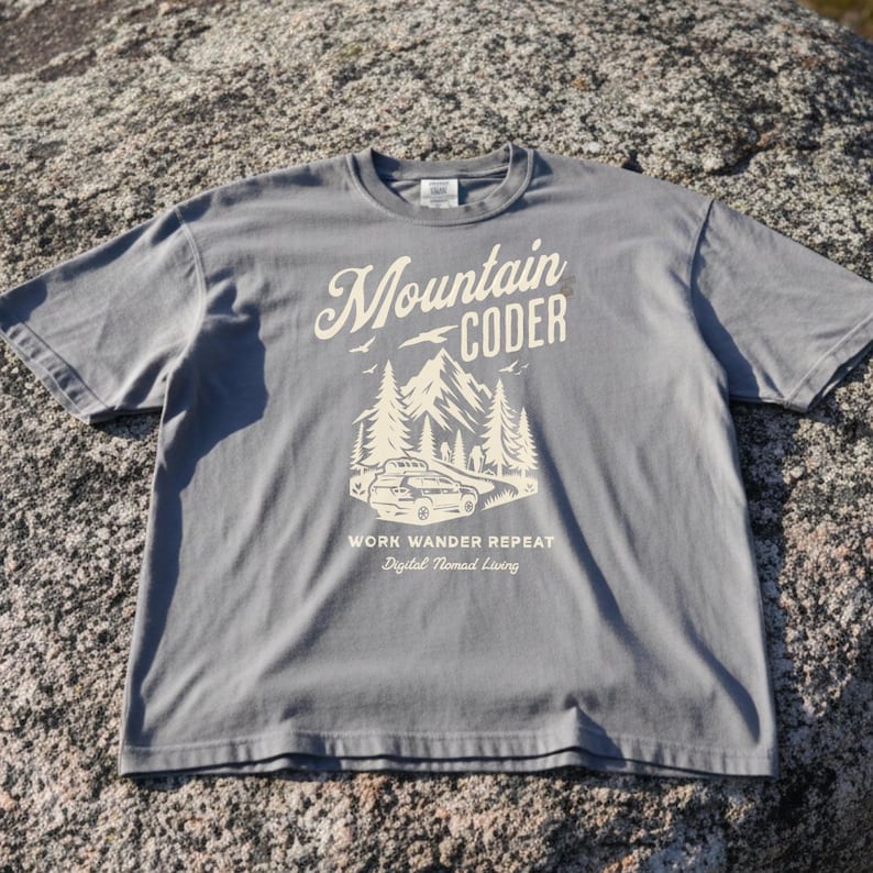Może przedstawiać: Szara koszulka z nadrukiem graficznym. Projekt zawiera słowa "Mountain Coder" nad sceną g&oacute;rską i leśną. Poniżej widnieją słowa "Work Wander Repeat" i "Digital Nomad Living".