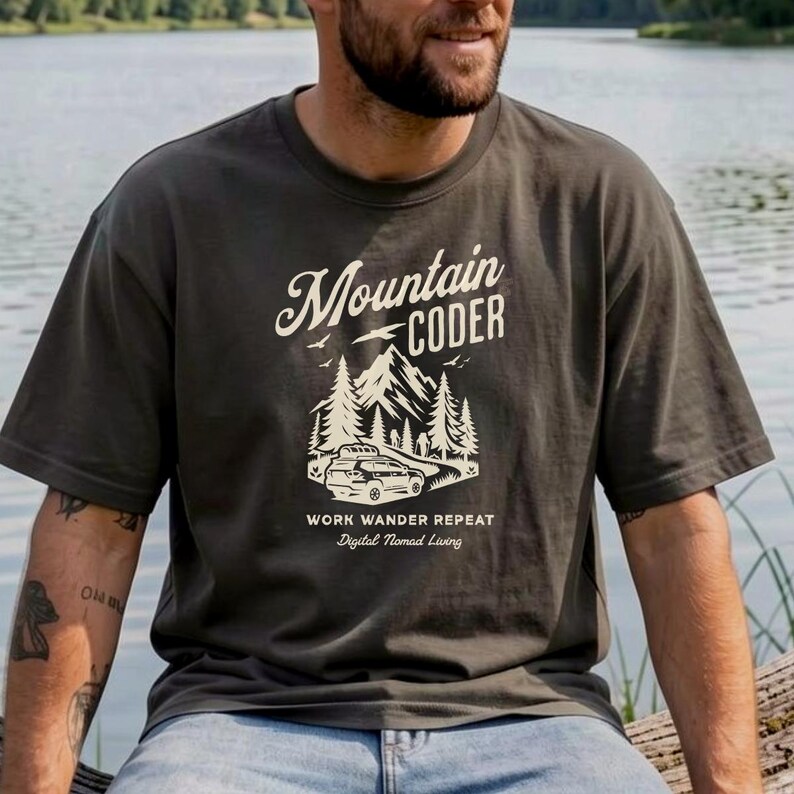 Może przedstawiać: Ciemnoszara koszulka z białym nadrukiem &bdquo;Mountain Coder&rdquo; nad ilustracją g&oacute;ry i samochodu. Poniżej grafiki znajduje się tekst &bdquo;Work Wander Repeat&rdquo; i &bdquo;Digital Nomad Living&rdquo;. Koszulka ma kr&oacute;tkie rękawy.
