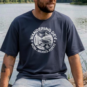 May include: Navy blue t-shirt featuring a white circular graphic with the text "Wandering Dev" and a mountain and lake scene. The words "Work-Wander-Repeat" are also included. The shirt is short-sleeved and made of a soft fabric.