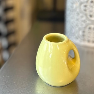 May include: A small, yellow ceramic vase with a rounded body and a handle. The vase has a smooth, glossy finish and a circular opening at the top. The handle is a loop shape. The vase is sitting on a metallic surface.