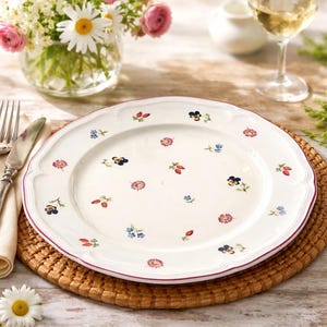 May include: White ceramic plate with a scalloped edge and a red trim, decorated with small floral designs in various colors. The plate is set on a woven placemat, with a fork, knife, and napkin to the left.