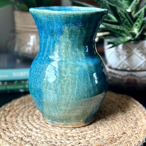 May include: A handcrafted ceramic vase with a glossy, turquoise glaze. The vase has a wide body and a flared rim, sitting on a round, woven jute mat. The background includes potted plants.