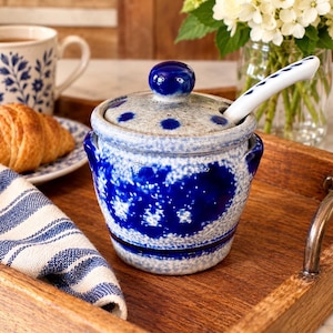 May include: A ceramic sugar bowl with a speckled light gray and deep blue glaze, featuring a matching lid with a knob handle and a small white ceramic spoon with blue leaf patterns. The bowl has two side handles and a rounded, sturdy base.