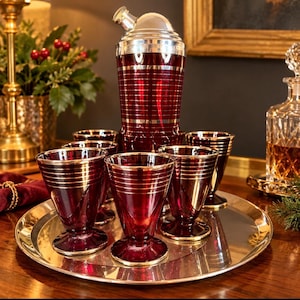 May include: A cocktail shaker and six matching glasses made of deep red glass with horizontal silver bands. The shaker features a silver metal lid and cap. The glasses are footed with a tapered shape, all arranged on a round silver metal tray.
