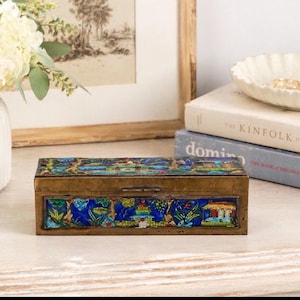 Puede incluir: Una caja rectangular de latón con un diseño de esmalte colorido. La caja presenta una escena de paisaje con acentos azules, verdes y amarillos. La caja está colocada sobre una superficie blanca, con libros y un cuenco decorativo en el fondo.