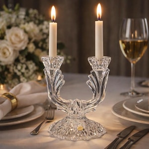 May include: A clear crystal double candlestick with two lit white candles. The candlestick has a decorative, curved design and sits on a table set for a formal dinner. A wine glass and a bouquet of white flowers are in the background.